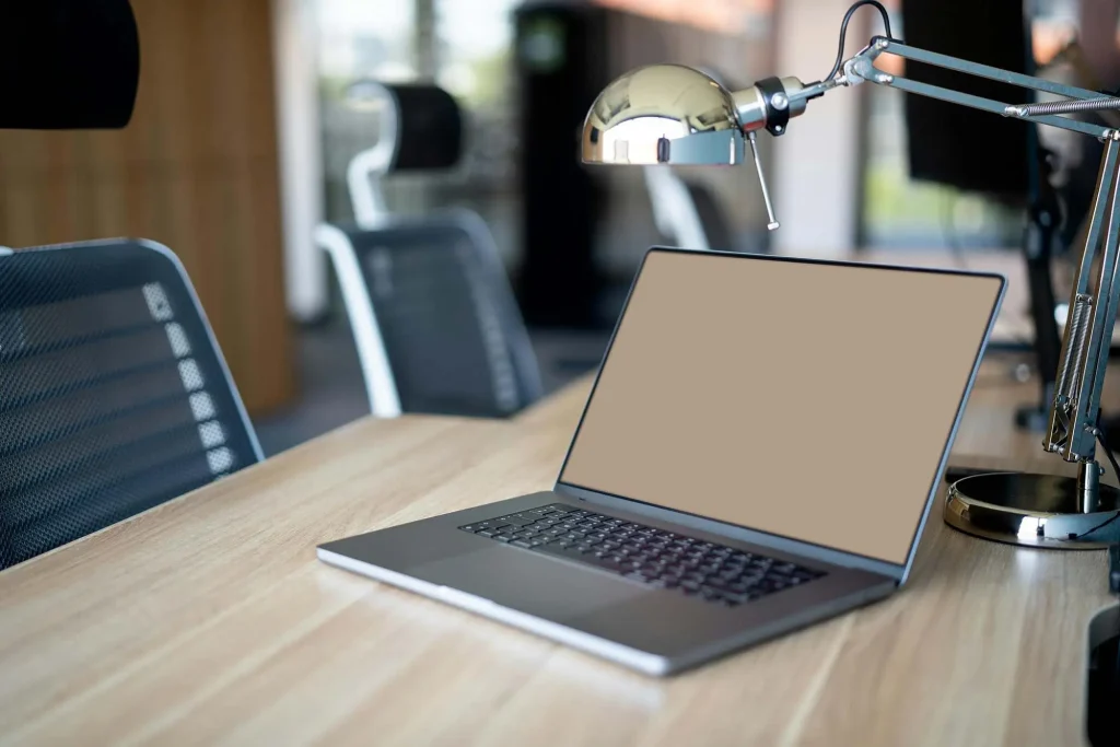 A laptop with a blank screen sits on a wooden desk beside a desk lamp in an office setting with empty chairs in the background, reminiscent of a well-organized Christchurch boudoir workspace.