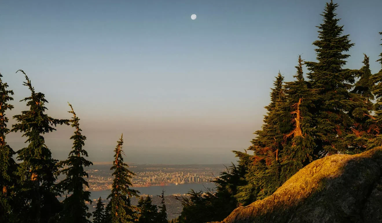 View from a rocky, forested hillside at sunset with the moon in the sky and a distant city visible on the horizon.