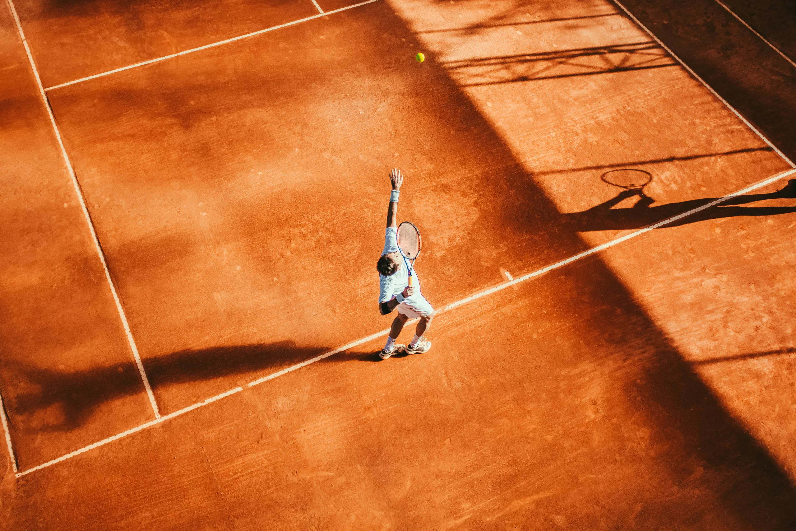 Tennis player in blue shirt serves on a clay court, casting a long shadow. The ball is in mid-air above the player's head.
