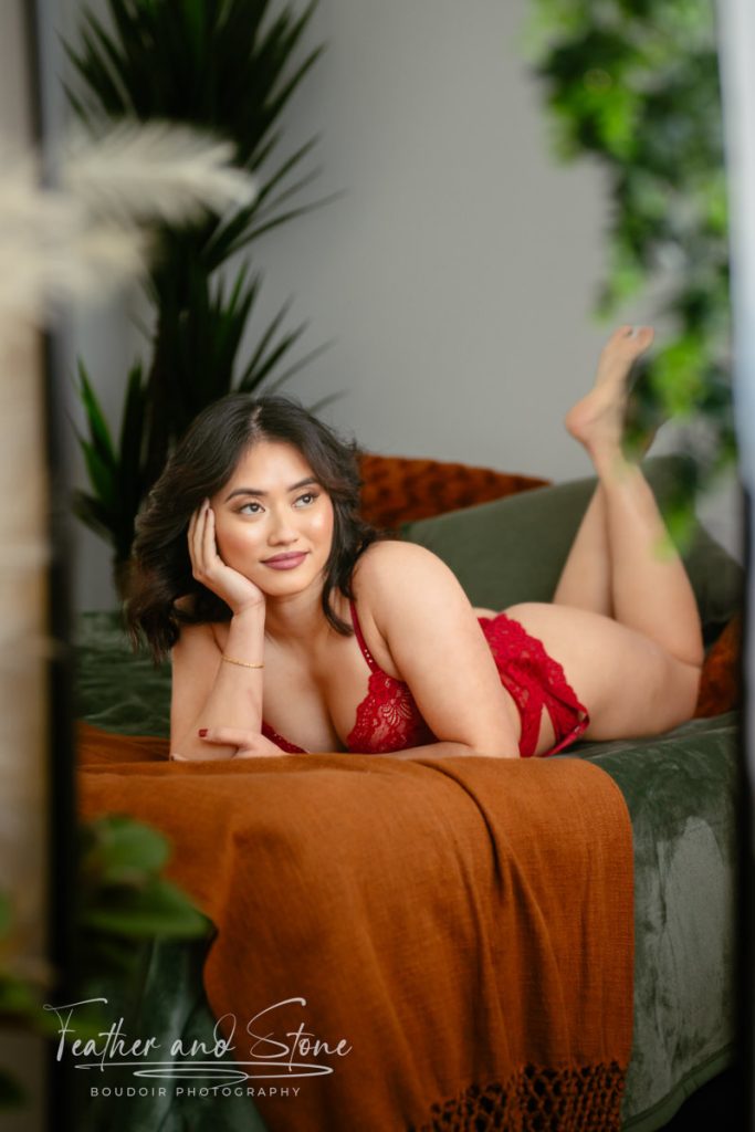 A person in red lace lingerie lies on a bed with an orange blanket, leaning on one arm, surrounded by green plants and muted lighting, like a scene from glowing customer reviews.