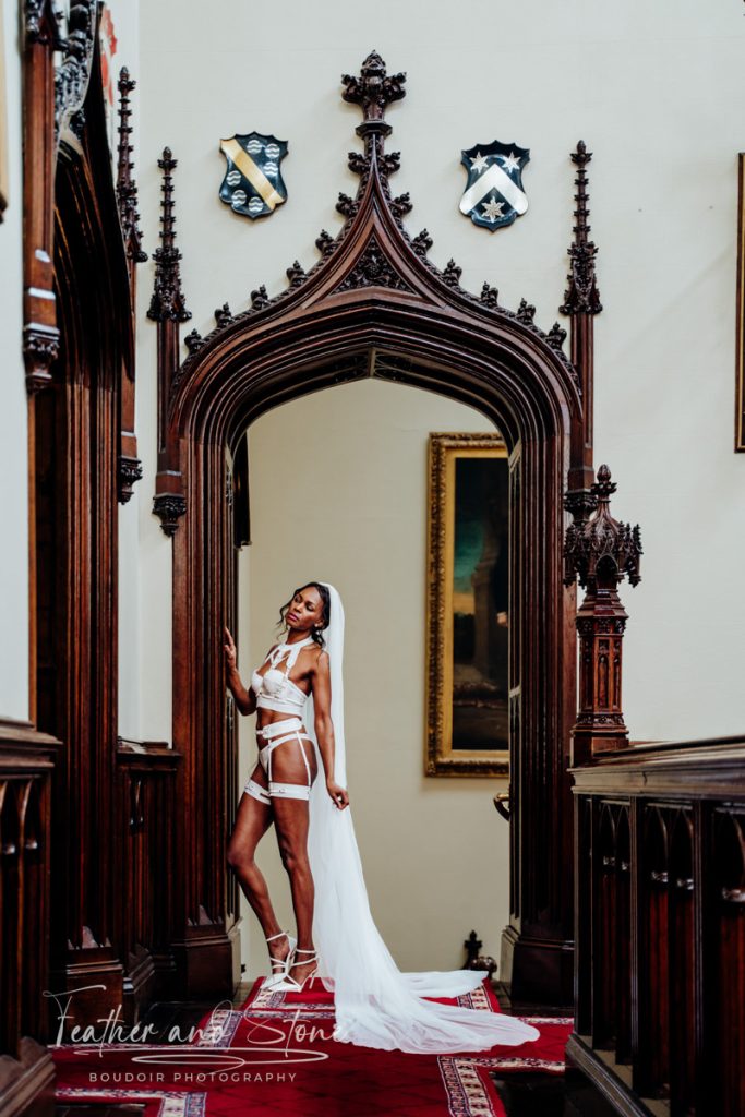 A woman in bridal lingerie and a veil poses in an ornate archway with wood carvings and shield decorations in the background. The scene, akin to the testimonials of elegance, is set in an elegant hall with rich decor.