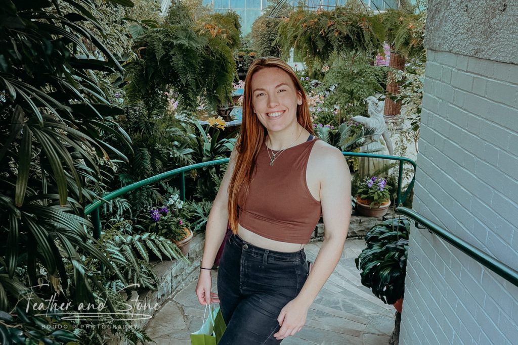 A person with long hair wearing a brown crop top and black jeans stands on a pathway surrounded by lush greenery and plants, providing a serene snapshot about nature.