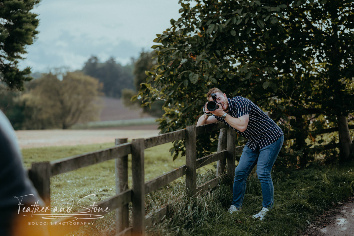 A photographer wearing a striped shirt and jeans leans on a wooden fence, holding a camera to their eye, capturing a shot in an outdoor, rural setting with trees and fields in the background. This scene is about the essence of nature's tranquility intertwined with artistic endeavor.