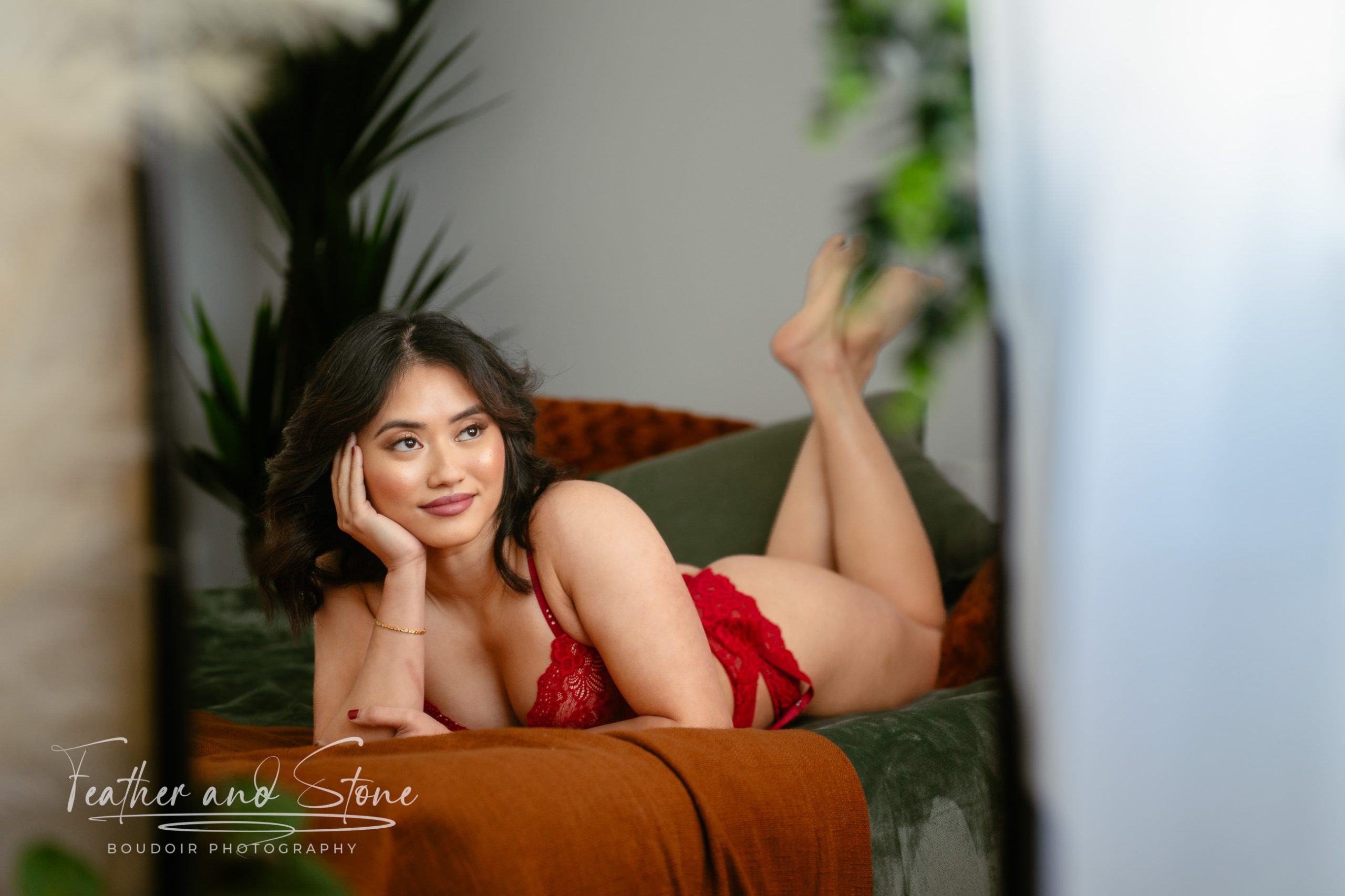 A woman in red lingerie lies on her stomach on a bed, propped up on her elbows, with one hand touching her hair. The background features green plants and pillows. "Feather and Stone Boudoir Photography" is visible.