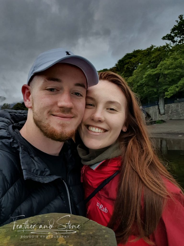 A man and a woman smile while taking a selfie outdoors. They are wearing jackets and standing in front of a body of water and trees. The sky is overcast, adding to the serene atmosphere about them.