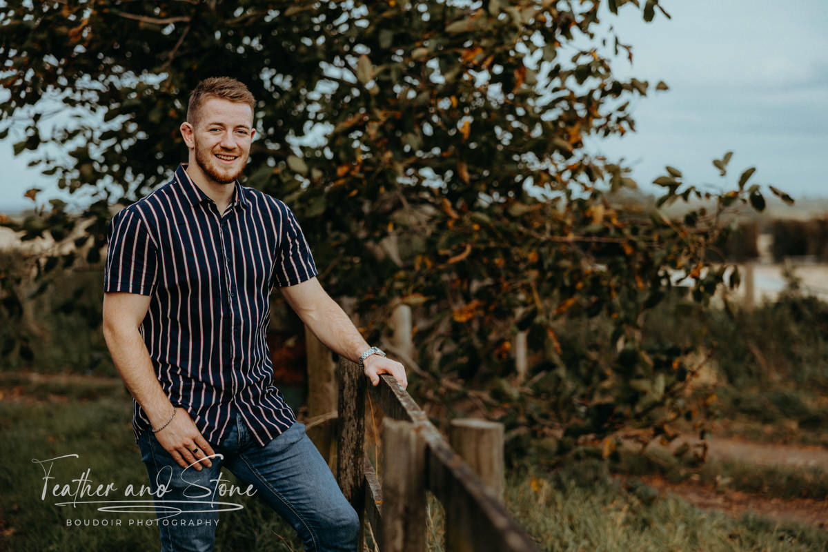A man in a striped shirt and jeans leans against a wooden fence outdoors, surrounded by trees and greenery, smiling at the camera. It appears to be about enjoying the simple moments in nature.