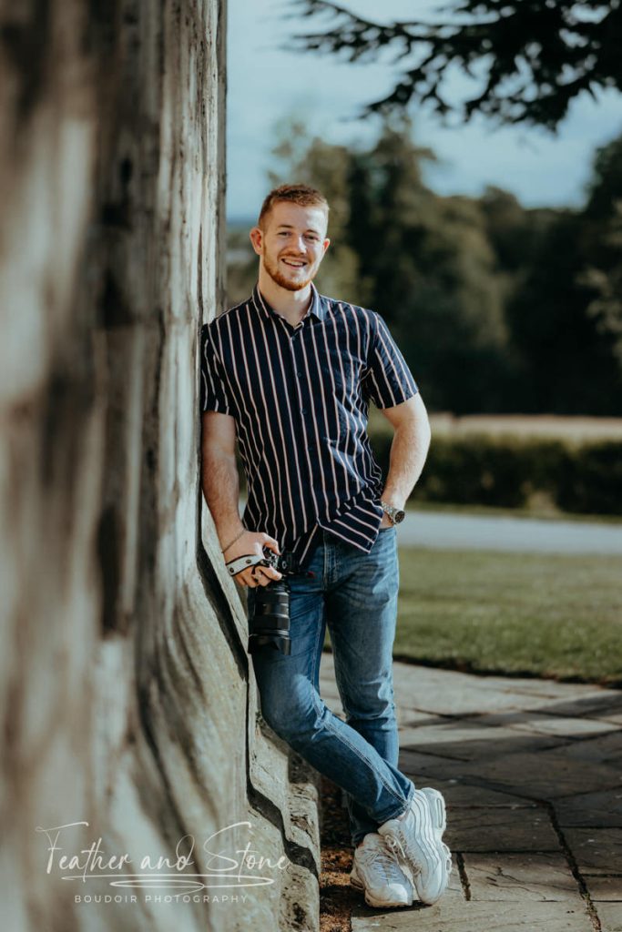 Feather and Stone boudoir Photographer, Niall Featherstone wearing a striped shirt and jeans leans against a stone wall outdoors, capturing the essence of their surroundings with a camera. Trees and a pathway adorn the background, evoking a sense of home.