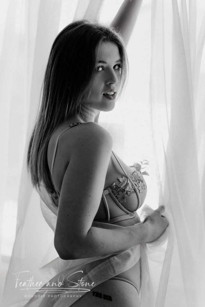 A woman in lingerie stands against a sheer curtain, looking at the camera. The photo is in black and white, with "Feather and Stone Boudoir Photography" text at the bottom.