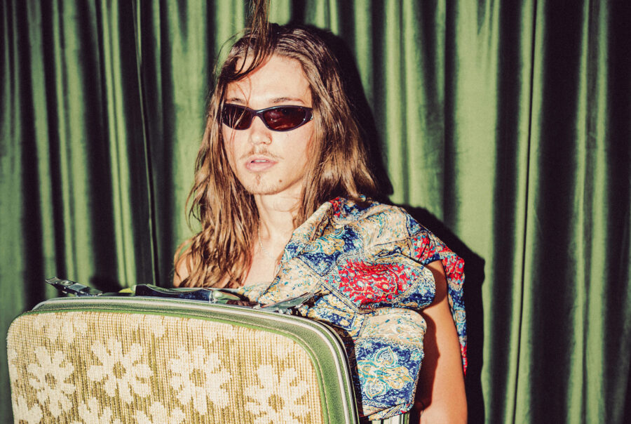 A person wearing sunglasses with long hair holds a floral-patterned suitcase and a colorful patterned garment, standing in front of green curtains.