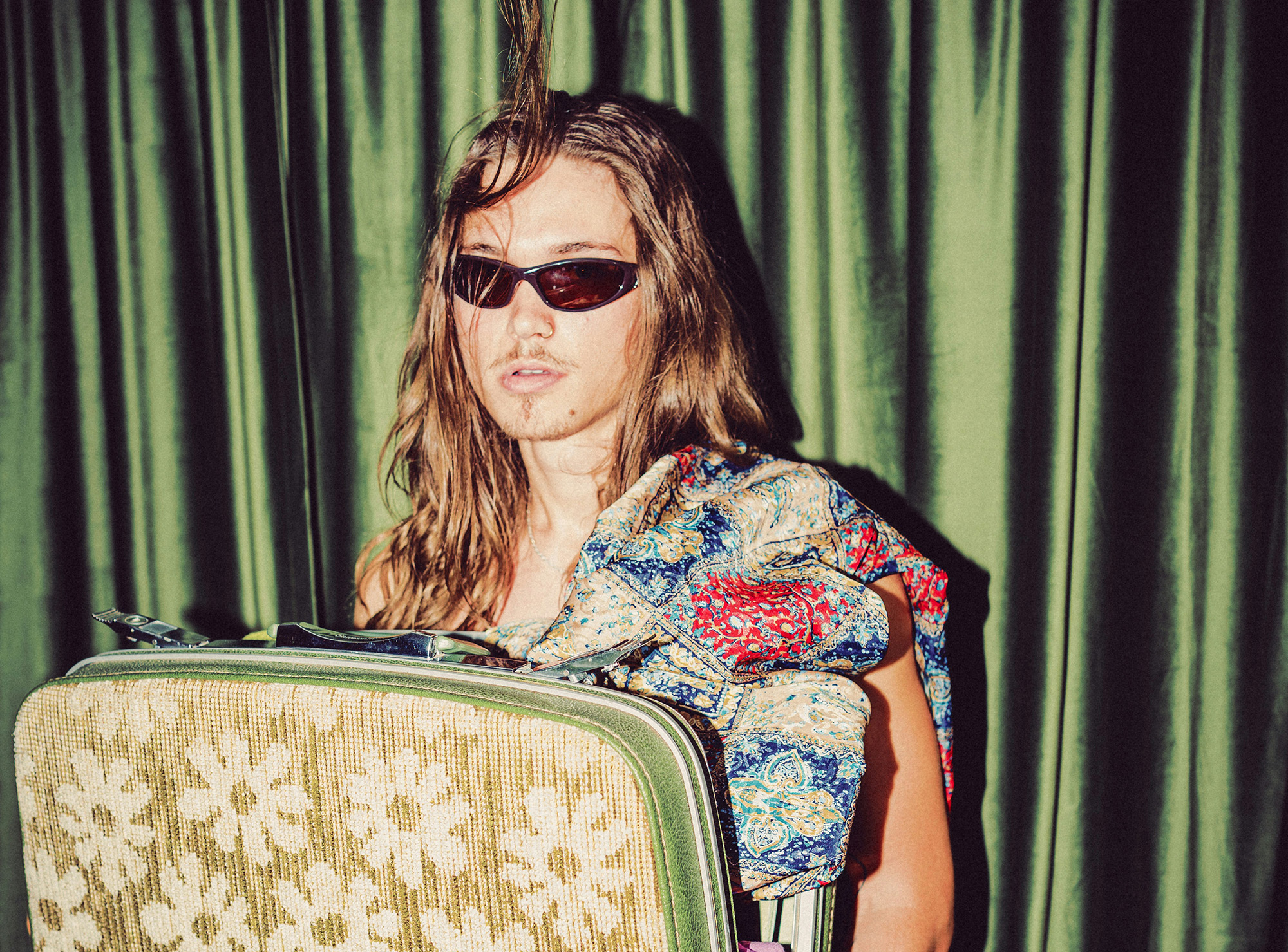 A person wearing sunglasses with long hair holds a floral-patterned suitcase and a colorful patterned garment, standing in front of green curtains.