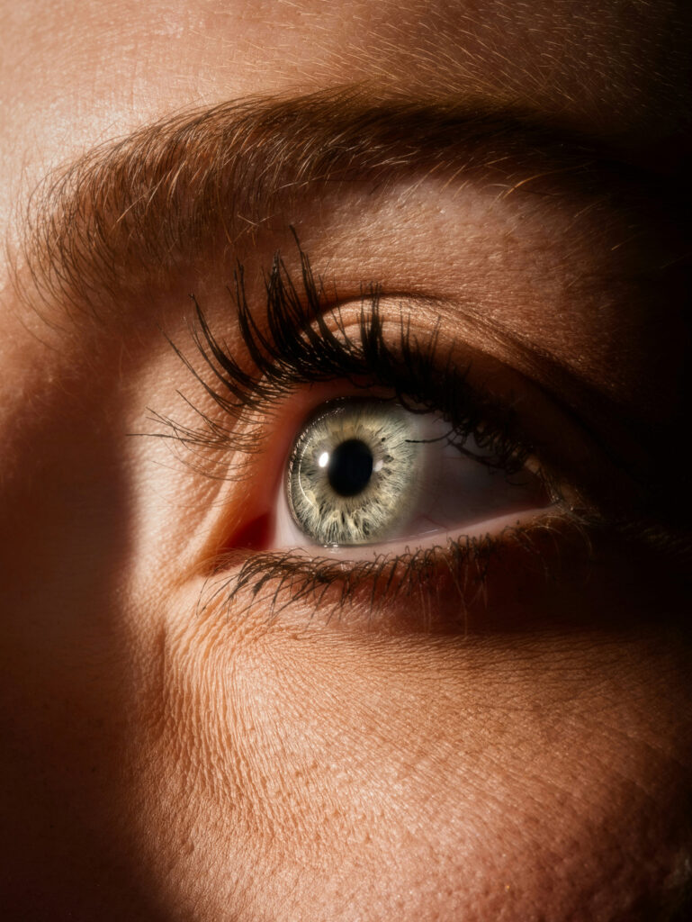 Close-up of a human eye with green iris, visible eyelashes, eyebrow, and skin texture in natural lighting.
