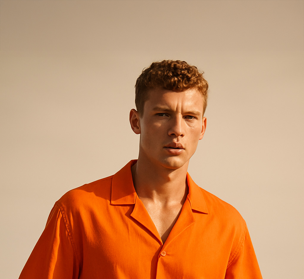 A young man with short curly hair wears an orange button-up shirt and stands against a plain beige background.