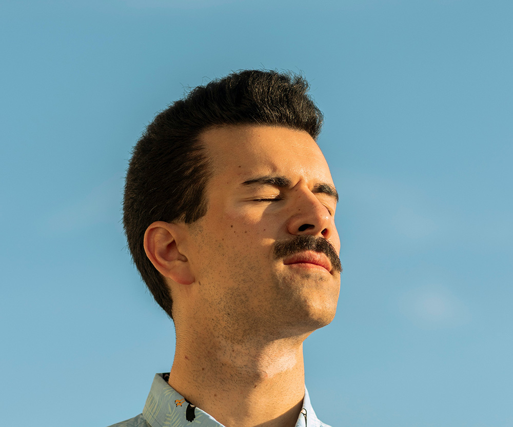 A man with a mustache stands outdoors with his eyes closed, facing the sun against a clear blue sky.