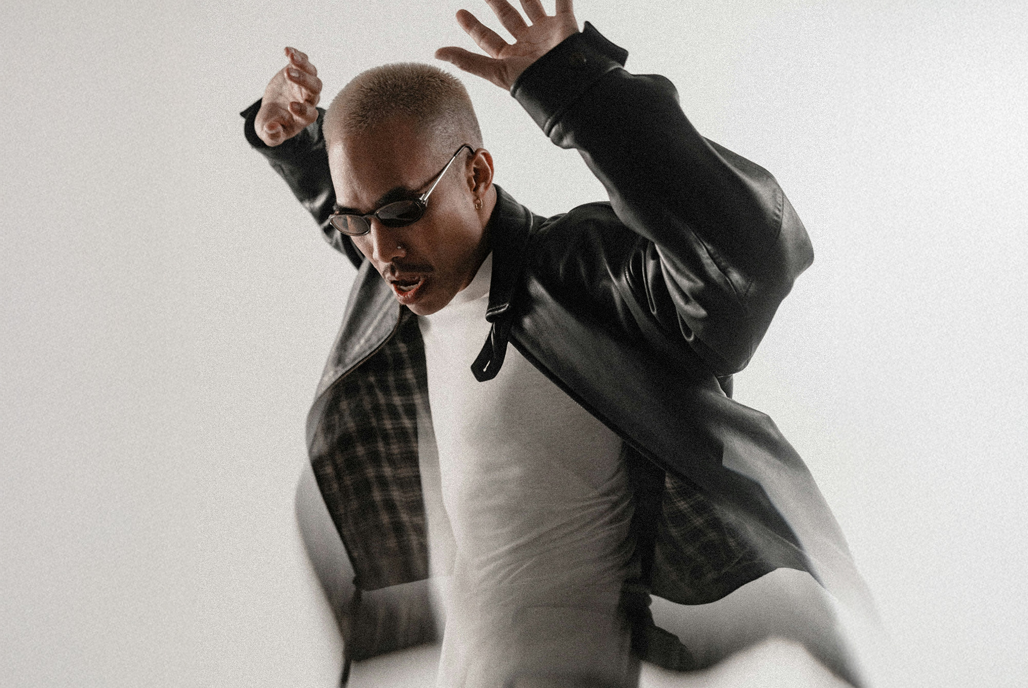 A person in sunglasses and a black leather jacket poses with arms raised against a plain background.