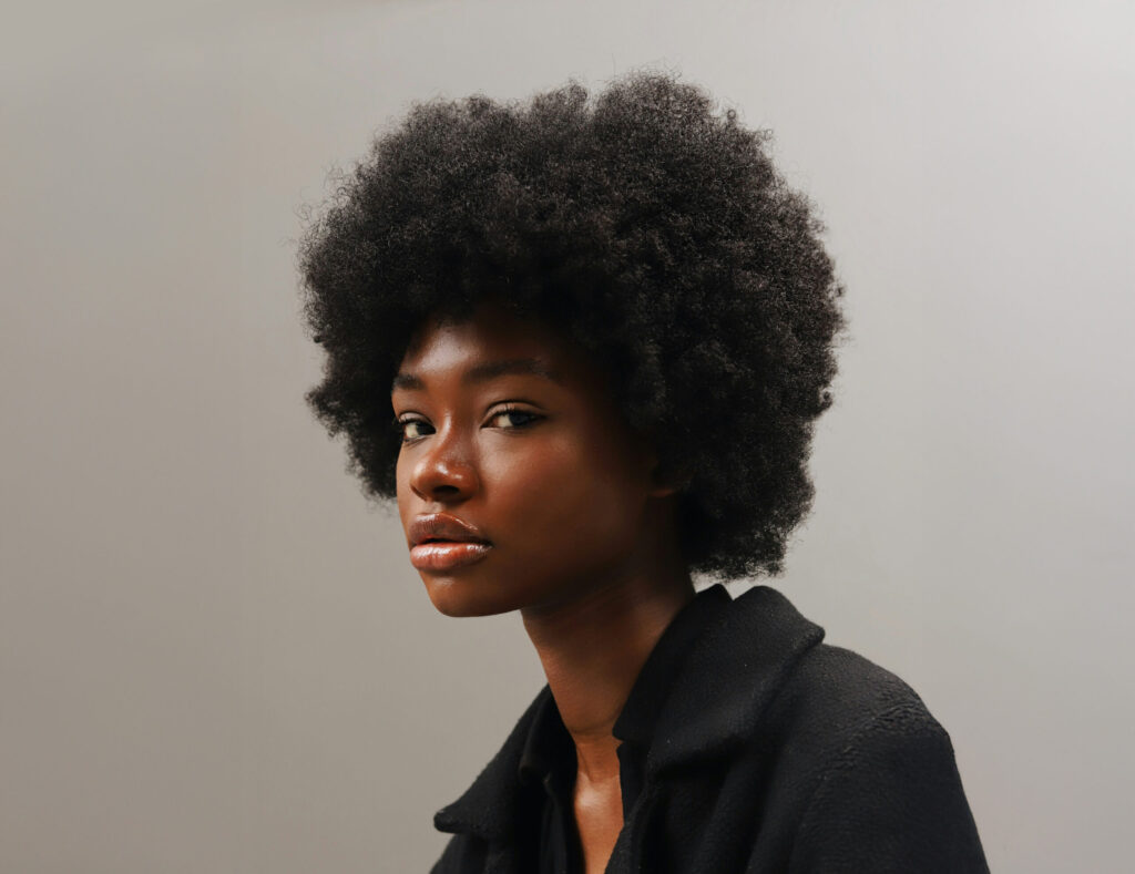A woman with a natural afro hairstyle wearing a black jacket looks at the camera against a plain, light background.