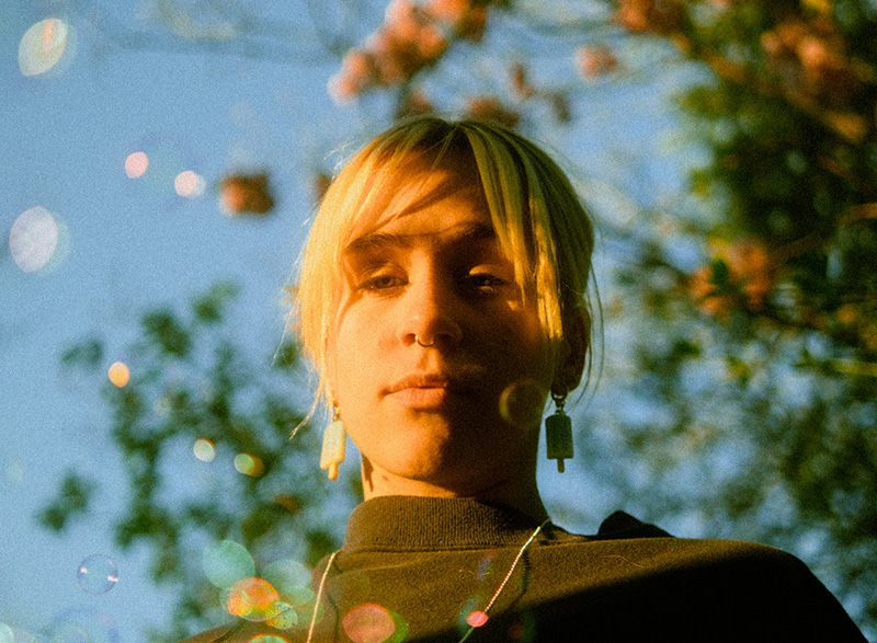 A person with short blonde hair and earrings stands outdoors, with sunlight and blurred foliage in the background.