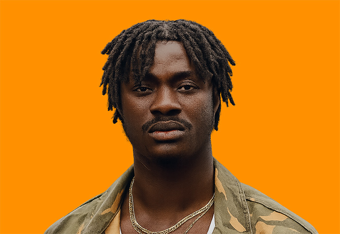 A man with short dreadlocks, wearing a camouflage jacket and gold chains, stands against a solid orange background.
